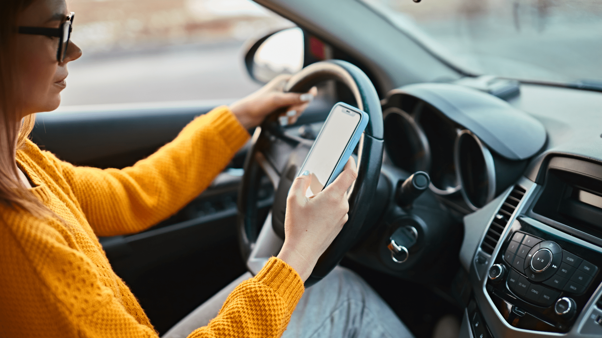 Woman in yellow jumper holding phone in right hand and steering wheel in left